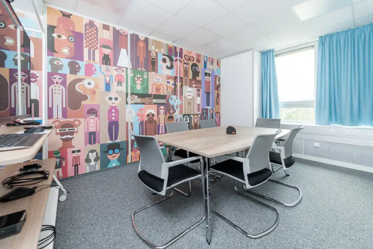 Büro mit farbenfrohem Wandgemälde, Tisch, Stühlen und einem Fenster mit blauen Vorhängen.