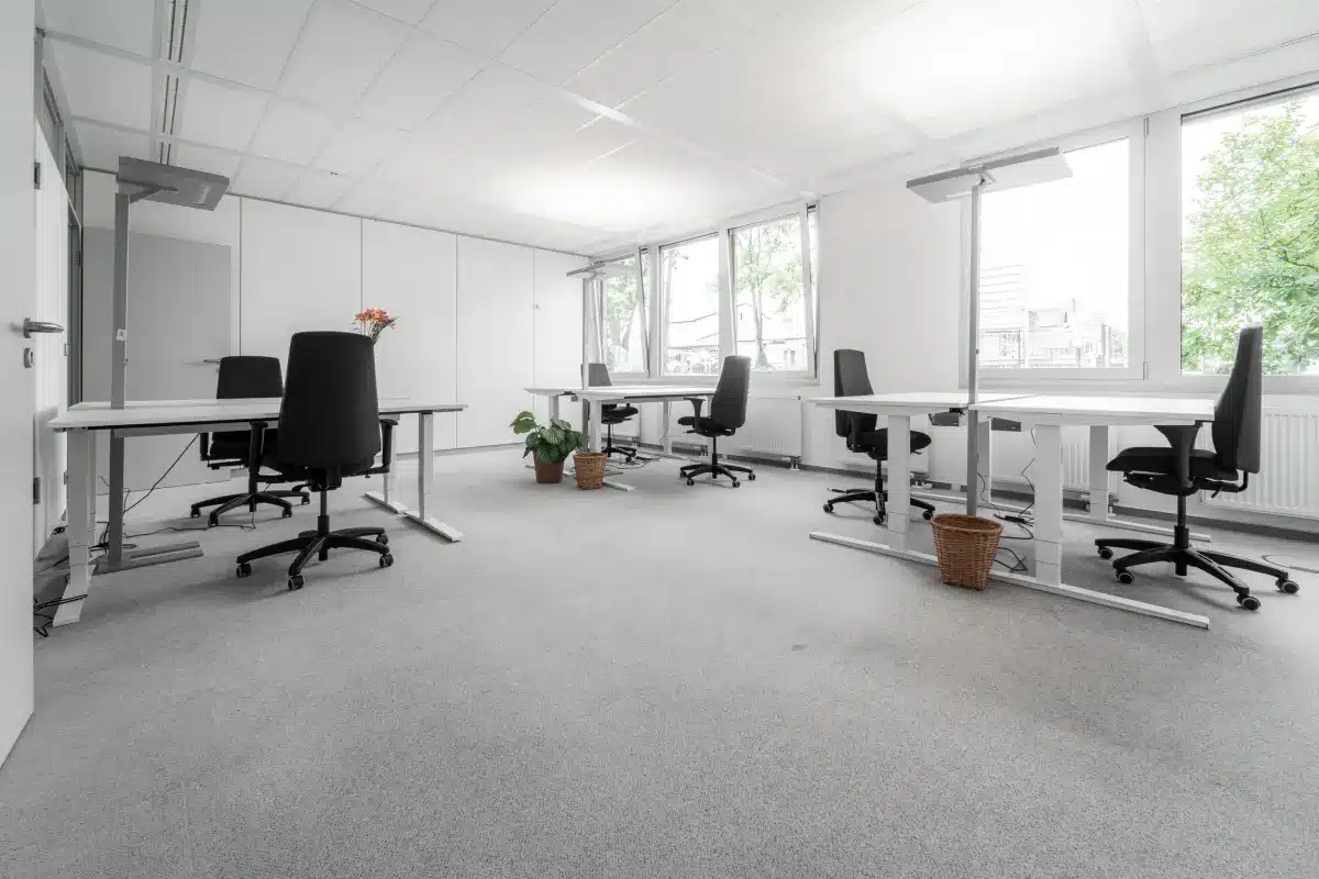 Geräumiges Büro mit Schreibtischen, Stühlen und Topfpflanzen, in der Nähe großer Fenster mit Tageslicht.